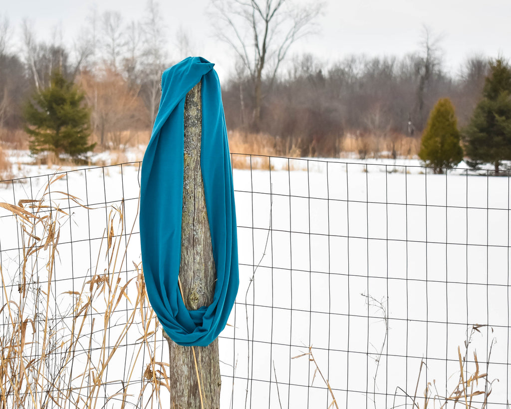 A teal blue infinity scarf hangs on a farm fence post with a snowy meadow in the backround