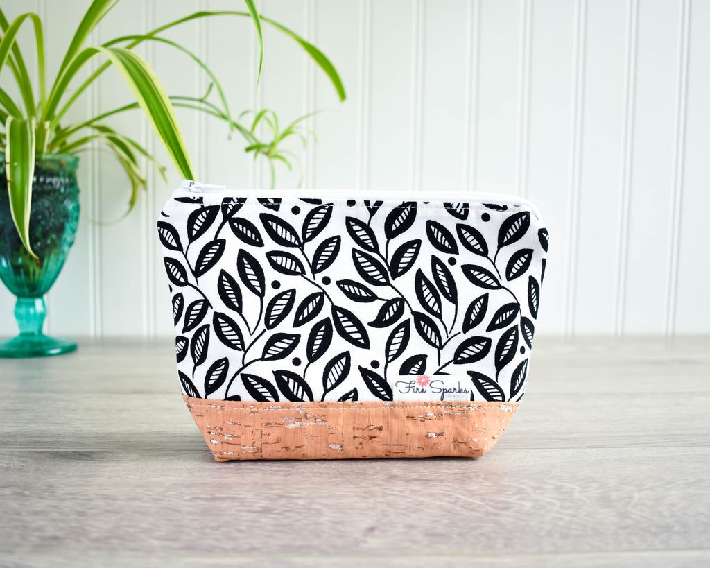 A zipped pouch with black and white leaf design and a cork base sits on a grey floor, white bead board in visible in the background along with a green plant in a tall green glass slightly out of focus on the left.