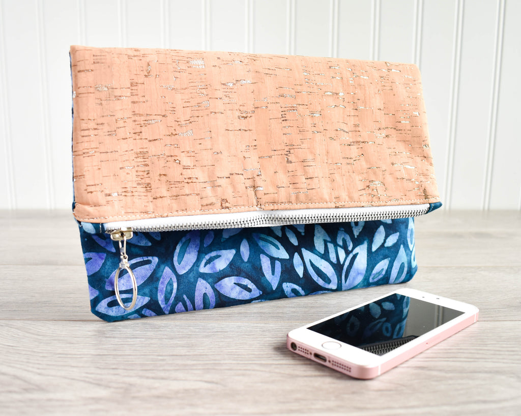 A foldover clutch with blue and purple batik flowers on one side and natural cork with silver flecks on the other sitting on a grey wooden floor with an iphone for scale