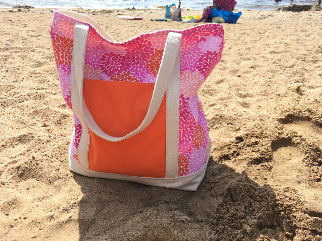 A large pink, orange, and canvas tote bag sits on a sunny, sandy beach.